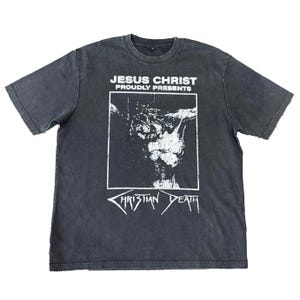 May include: Dark gray t-shirt featuring a graphic of Jesus Christ with the text "JESUS CHRIST PROUDLY PRESENTS" above the image and "CHRISTIAN DEATH" below. The shirt has a crew neck and short sleeves.