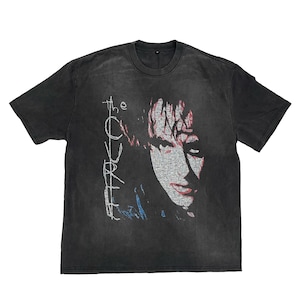 May include: Dark gray t-shirt featuring a portrait of a person with the text "The Cure" in white. The graphic is in red, white, and blue. The shirt has a crew neck and short sleeves.