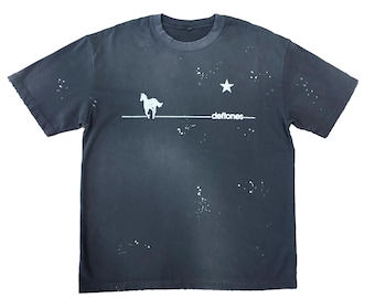May include: Dark gray t-shirt featuring a white horse graphic above the word "deftones" and a star. The shirt has a crew neck and short sleeves. White paint splatters are scattered across the shirt.