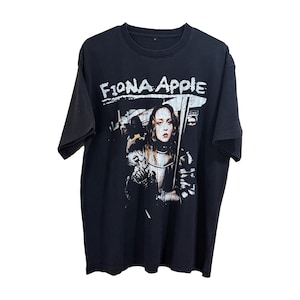 May include: Black t-shirt featuring the text "Fiona Apple" in white, above a graphic of a woman in armor holding a sword. The image has a distressed, vintage look.