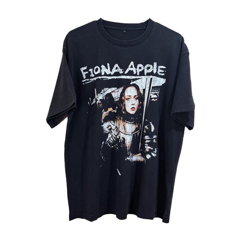 May include: Black t-shirt featuring the text "Fiona Apple" in white, above a graphic of a woman in armor holding a sword. The image has a distressed, vintage look.