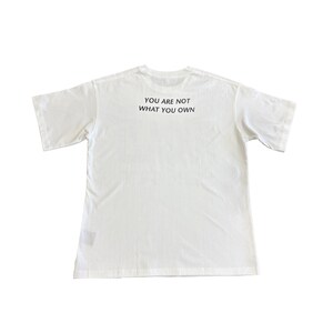 May include: A white t-shirt with short sleeves. The text "YOU ARE NOT WHAT YOU OWN" is printed in black on the back of the shirt. The shirt is made of a soft, comfortable material.