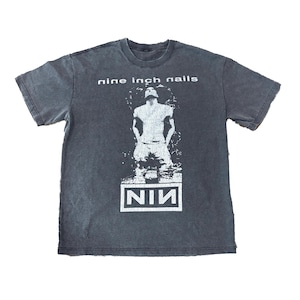 May include: Dark grey t-shirt with the words "nine inch nails" above a white graphic of a person and the band's logo "NIN". The shirt has a crew neck and short sleeves.