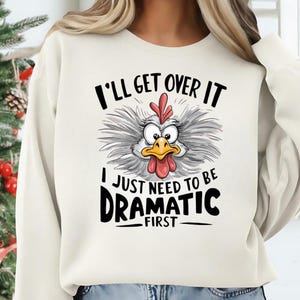 May include: Cream-colored sweatshirt featuring a cartoon chicken with the text "I'll Get Over It, I Just Need To Be Dramatic First." The chicken has a red comb and wattle, with a yellow beak and wide eyes. The text is in black.