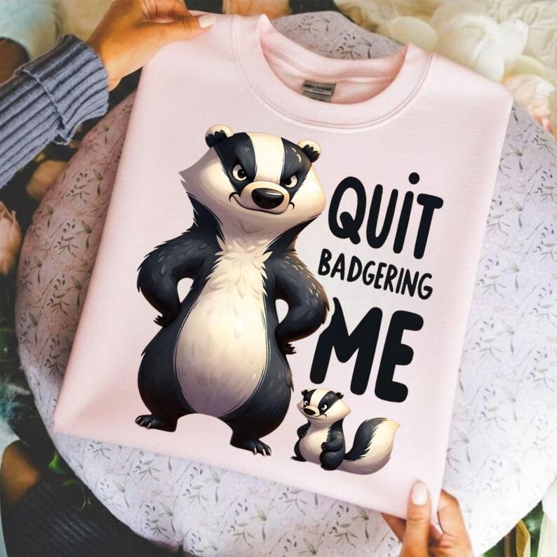 May include: Light pink sweatshirt featuring a cartoon badger with the text "Quit Badgering Me." The badger is black, white, and tan. A smaller badger is also featured. The sweatshirt has a crew neck.