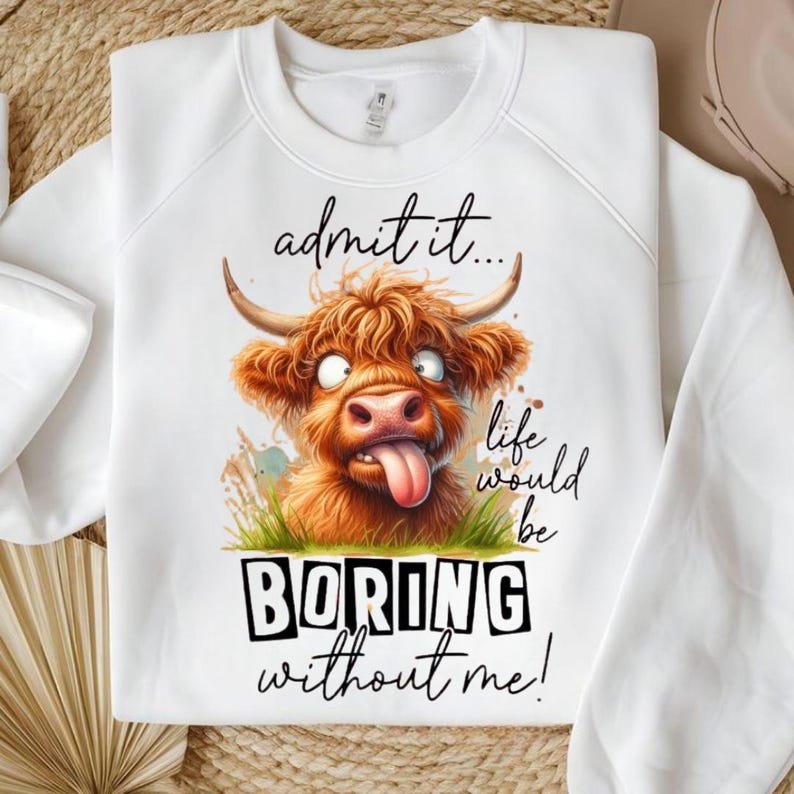 May include: White sweatshirt featuring a cartoon Highland cow with a tongue sticking out. The cow is brown with horns and is set against a background of green grass and the words "admit it... life would be BORING without me!"