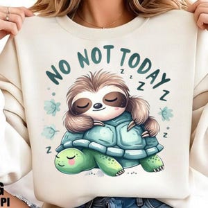 May include: Cream-colored sweatshirt featuring a cartoon sloth sleeping on a turtle. The sloth is brown and tan, the turtle is green and blue. The text "NO NOT TODAY" is above the sloth, with "Z"s around the image.