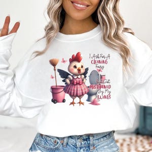 May include: White long-sleeve sweatshirt with a whimsical graphic. The graphic features a cartoon chicken with wings, a pink plaid dress, and the text "I Ask For A Cleaning Fairy But Got Mistaken By My Wings."