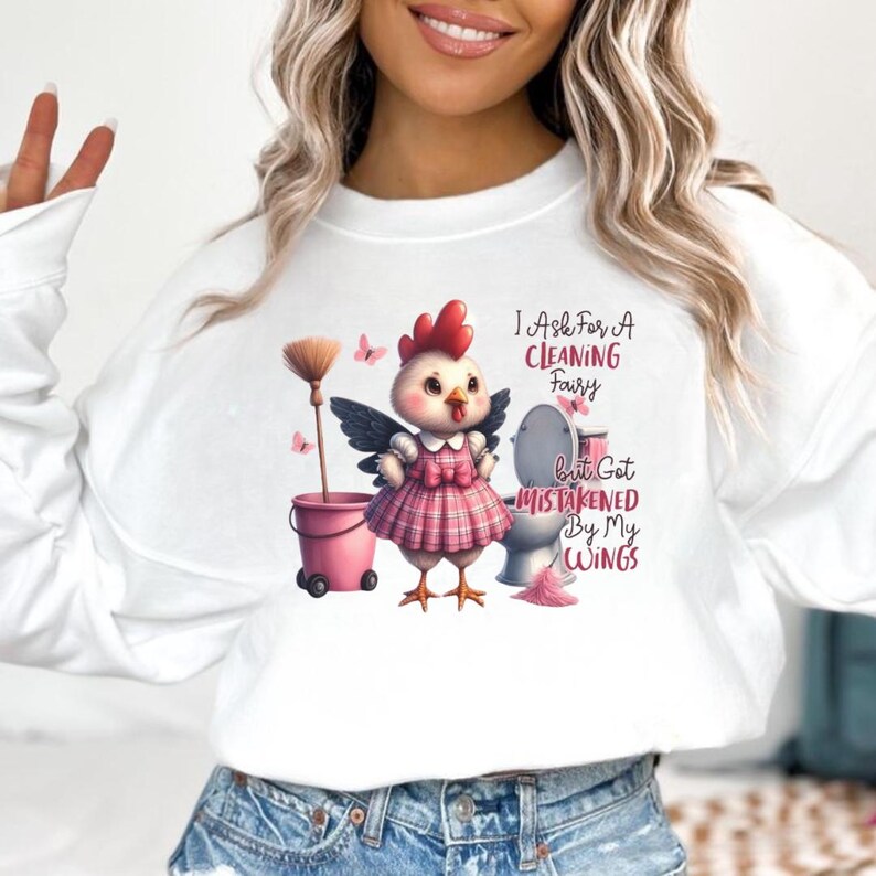 May include: White long-sleeve sweatshirt with a whimsical graphic. The graphic features a cartoon chicken with wings, a pink plaid dress, and the text "I Ask For A Cleaning Fairy But Got Mistaken By My Wings."