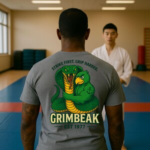 May include: A gray t-shirt with a graphic of a green cobra and a green bird hugging. The text "STRIKE FIRST. GRIP HARDER." arches above the graphic. Below the graphic is the word "GRIMBEAK" and "EST 1977".