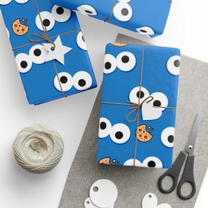 Op de afbeelding: Blauw cadeaupapier met een herhalend patroon van cartoonogen en koekjes. De ingepakte cadeaus zijn vastgebonden met touw en versierd met witte labels. Een bol touw, een schaar en extra labels zijn ook zichtbaar.