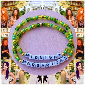 May include: Two beaded bracelets with green and gold beads. The bracelets have white letter beads spelling "MIDNIGHT MARGARITAS". The bracelets are on a pink surface, with images of the movie "Practical Magic" in the background.