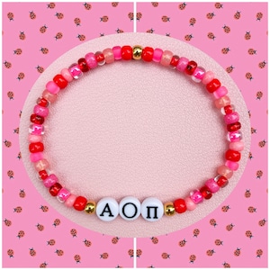 May include: A beaded bracelet in shades of red and pink, with gold accents and white letter beads spelling out "A O Π". The bracelet is displayed on a pink surface with a pink background decorated with ladybug patterns. A colorful accessory.