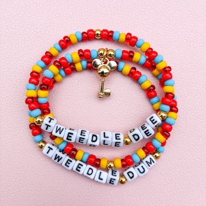 May include: Two beaded bracelets featuring red, yellow, and light blue beads with gold-tone accents. One bracelet displays the white letter beads spelling TWEEDLE DEE, while the other spells TWEEDLE DUM. A gold-tone key charm hangs from the top bracelet.