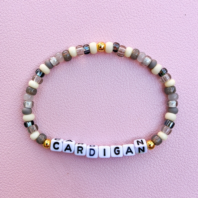 May include: A beaded bracelet featuring a repeating pattern of cream, translucent grey, and metallic gold beads. The center of the bracelet displays white square letter beads that spell out the word CARDIGAN in black capital letters.