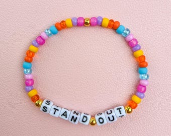 May include: A colorful beaded bracelet with the word "STAND OUT" spelled out in white letter beads. The bracelet features a variety of beads in shades of pink, orange, yellow, blue, and purple, with gold accents. The bracelet is on a pink surface.