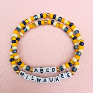 May include: Two circular beaded bracelets featuring a repeating pattern of yellow, cream, navy blue, and metallic gold beads. One bracelet includes white square beads with black letters spelling out ABCD, and the other spells out MILWAUKEE.
