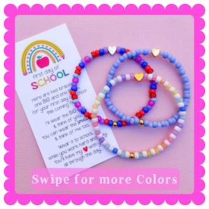 May include: Two beaded bracelets featuring small colorful glass beads in shades of red, blue, purple, white, and yellow, each accented with a small gold-tone heart charm. A white card is included with a rainbow graphic and the text first day of SCHOOL.