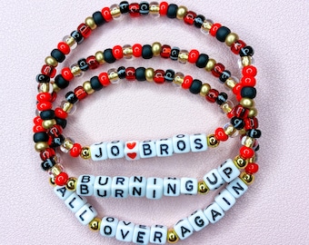 May include: Three elastic bracelets featuring red, black, and gold-toned beads. The bracelets include white square letter beads spelling out JO BROS with a red heart, BURNING UP, and ALL OVER AGAIN, arranged in a circular pattern on a light surface.
