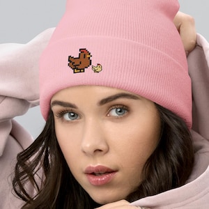 May include: A light pink beanie featuring a pixelated brown chicken and yellow chick design. The beanie is made of a soft material and is a comfortable accessory. The beanie is a great addition to any wardrobe.