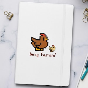 Op de afbeelding: Wit notitieboek met een gepixeld bruin kippen- en kuikendesign, met de tekst "busy farmin'". Het notitieboek ligt op een wit oppervlak met een toetsenbord, koffiekopje en potloden.