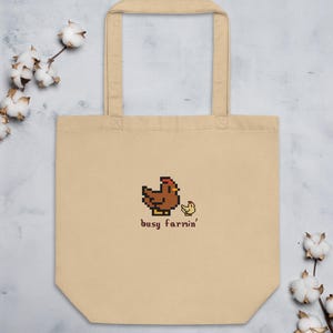 Op de afbeelding: Beige draagtas met een gepixeld bruin kippen- en geel kuikendesign. De tekst "busy farrin'" staat onder de afbeelding. De tas heeft twee handvatten en is gemaakt van een lichtgekleurde stof.