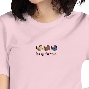 Puede incluir: Camiseta rosa claro con un diseño pixelado de tres pollos amarillos, marrones y azules. El texto "busy farnin'" está debajo. La camiseta está hecha de un material suave.