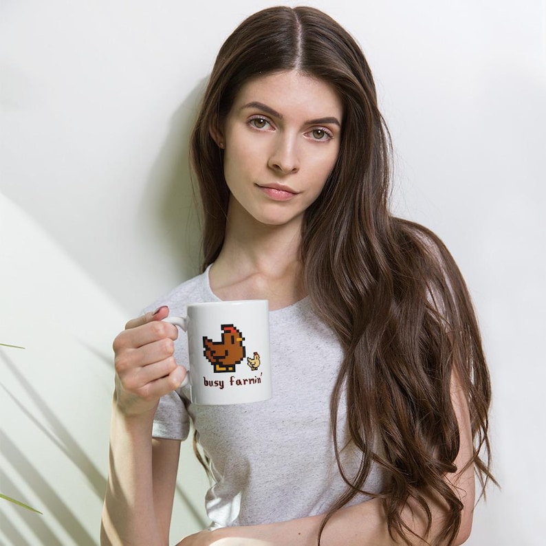 Puede incluir: Taza de cer&aacute;mica blanca con un pollo y un pollito marrones pixelados y el texto "busy farrin'". La taza la sostiene una persona que lleva una camiseta gris.