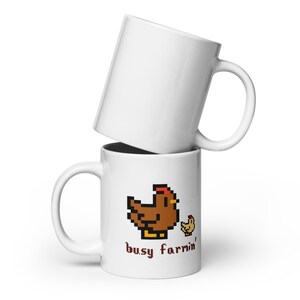 Puede incluir: Dos tazas de cer&aacute;mica blanca apiladas. La taza inferior presenta un dise&ntilde;o pixelado de gallina y pollito marr&oacute;n con el texto "busy farmin'". La taza superior es de color blanco liso.