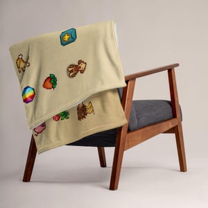 Op de afbeelding: Een beige sprei met pixelafbeeldingen van diverse objecten, gedrapeerd over een houten stoel. De sprei heeft een regenboog, een aardbei, een varken, een bij en een schatkist. De stoel heeft een grijze zitting en een houten frame.