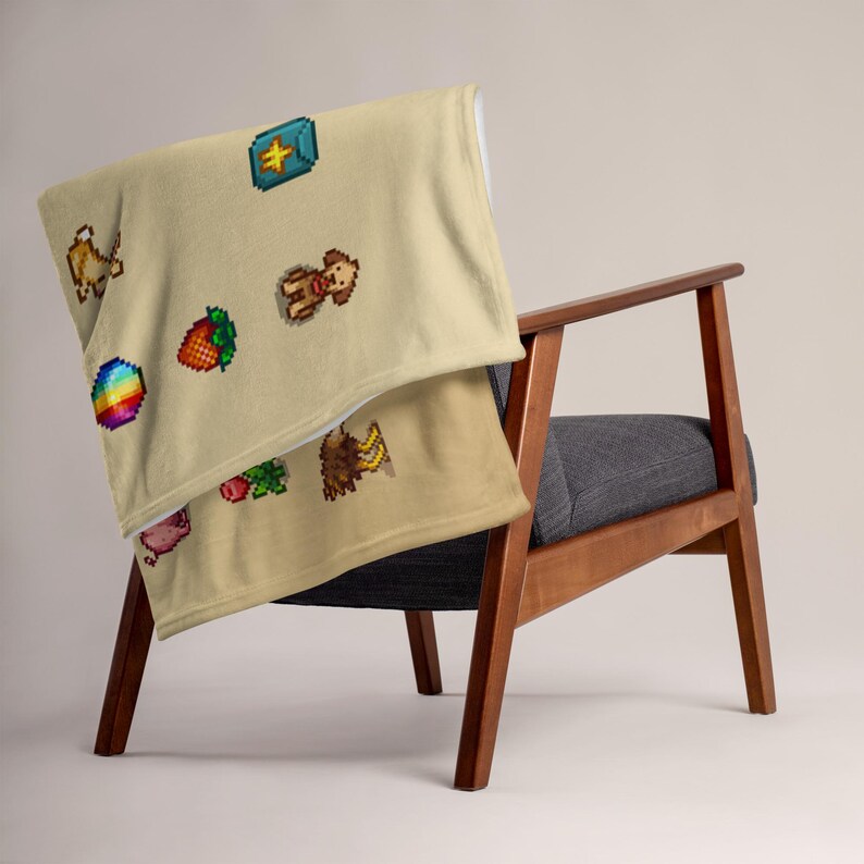 Op de afbeelding: Een beige sprei met pixelafbeeldingen van diverse objecten, gedrapeerd over een houten stoel. De sprei heeft een regenboog, een aardbei, een varken, een bij en een schatkist. De stoel heeft een grijze zitting en een houten frame.