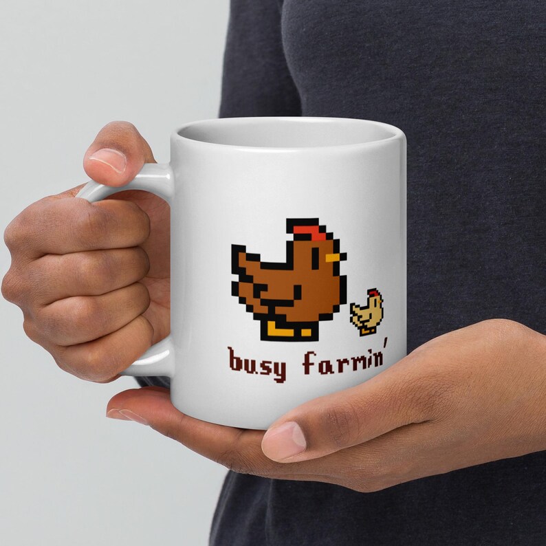 Puede incluir: Taza de cer&aacute;mica blanca con un dise&ntilde;o pixelado de gallina y pollito marrones, con detalles rojos y amarillos. La taza tiene el texto "busy farmin'" en una fuente estilizada. La taza est&aacute; sujeta por una persona.