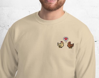 Puede incluir: Sudadera beige con pollos bordados pixelados y un bocadillo en forma de corazón. El diseño incluye un pollo amarillo y otro marrón, con un corazón rojo encima.