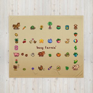 Op de afbeelding: Een beige deken met pixelafbeeldingen van boerderijgerelateerde items, waaronder een paard, wortel, schildpad, zonnebloem en diverse groenten en fruit. De zin "busy farmin'" is ook afgebeeld.