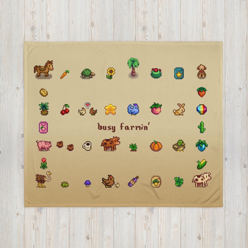 Op de afbeelding: Een beige deken met pixelafbeeldingen van boerderijgerelateerde items, waaronder een paard, wortel, schildpad, zonnebloem en diverse groenten en fruit. De zin "busy farmin'" is ook afgebeeld.