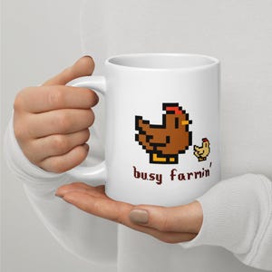 Puede incluir: Taza de cer&aacute;mica blanca con un dise&ntilde;o pixelado de gallina y pollito marrones y el texto "busy farmin'" en marr&oacute;n. La taza tiene un asa curva y la sostiene una persona.