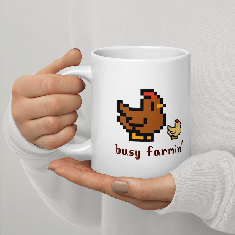Puede incluir: Taza de cer&aacute;mica blanca con un dise&ntilde;o pixelado de gallina y pollito marrones y el texto "busy farmin'" en marr&oacute;n. La taza tiene un asa curva y la sostiene una persona.