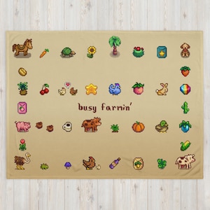 Op de afbeelding: Een beige deken met pixelafbeeldingen van boerderijdieren, gewassen en andere boerderijgerelateerde items. De deken heeft de tekst "busy farmin'" in het midden. De deken is gemaakt van een zacht materiaal.