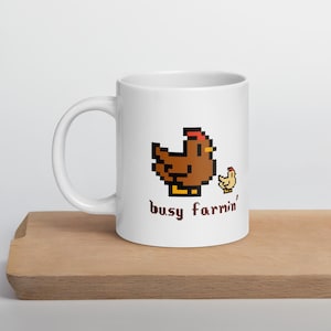 Pode incluir: Caneca de cerâmica branca com um desenho pixelizado de galinha e pintinho castanhos com o texto "busy farmin'". A caneca está sobre uma superfície de madeira.