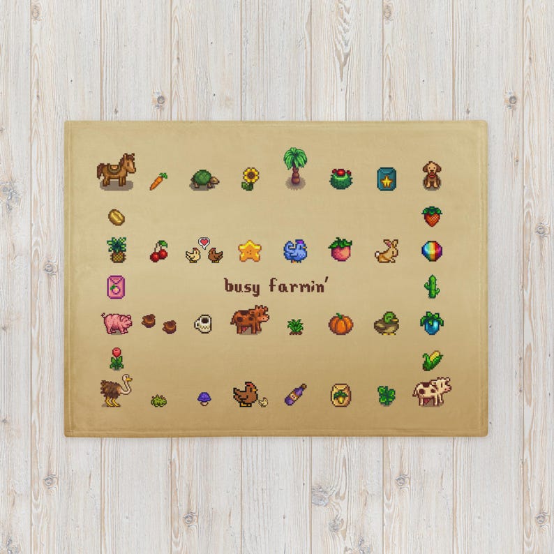 Op de afbeelding: Een beige deken met gepixelde illustraties van boerderijdieren, gewassen en andere boerderijgerelateerde iconen. De zin "busy farmin'" staat in het midden gedrukt. De deken is rechthoekig en lijkt van een zacht materiaal te zijn.