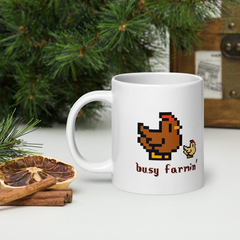 Puede incluir: Taza de cer&aacute;mica blanca con un dise&ntilde;o pixelado de gallina y pollito marrones y el texto "busy farrin'". La taza tiene un asa curva. El fondo incluye vegetaci&oacute;n y un cofre de madera.