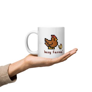 Puede incluir: Taza de cer&aacute;mica blanca con un dise&ntilde;o pixelado de gallina y pollito marrones y el texto "busy farrin'". La taza tiene un asa curva. El dise&ntilde;o es en marr&oacute;n, amarillo y rojo. La taza se sostiene en una mano.