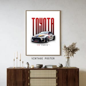 May include: A framed vintage poster featuring a white and black Toyota GR Yaris race car. The word "TOYOTA" is in red block letters above the car, with "- GR YARIS -" below. The poster is displayed on a wooden cabinet.