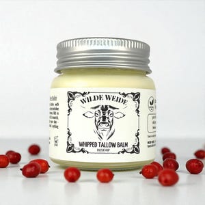 Puede incluir: Un pequeño tarro de cristal con tapa plateada, lleno de un bálsamo amarillo cremoso. La etiqueta dice "WILDE WEIDE" con una ilustración de vaca y "WHIPPED TALLOW BALM ROSEHIP". Varias escaramujos rojos están esparcidos alrededor del tarro.
