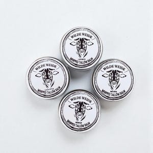 Puede incluir: Cuatro botes redondos blancos de Wilde Weide Whipped Tallow Balm, cada uno con una ilustración de vaca en blanco y negro. Los botes están etiquetados con diferentes aromas: Lavanda, Eucalipto, Natural y Escaramujo.