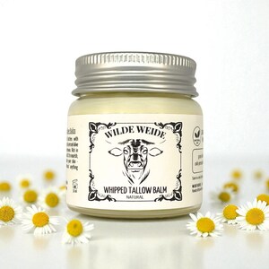 Könnte beinhalten: Ein Glas Wilde Weide Whipped Tallow Balm mit silbernem Deckel. Das Etikett zeigt eine Kuhillustration und die Worte "Whipped Tallow Balm" und "Natural". Das Glas ist von kleinen weißen und gelben Blumen umgeben.