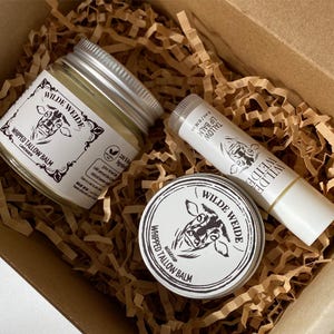 Könnte beinhalten: Eine Geschenkbox mit drei Wilde Weide-Produkten: ein Glas geschlagenes Talgbalsam, ein Lippenbalsam und eine kleine Dose geschlagenes Talgbalsam. Die Produkte zeigen eine Kuhillustration.