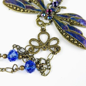 May include: A bronze dragonfly pendant with blue and purple iridescent wings, a decorative filigree charm, and two blue crystal beads hanging from a chain.