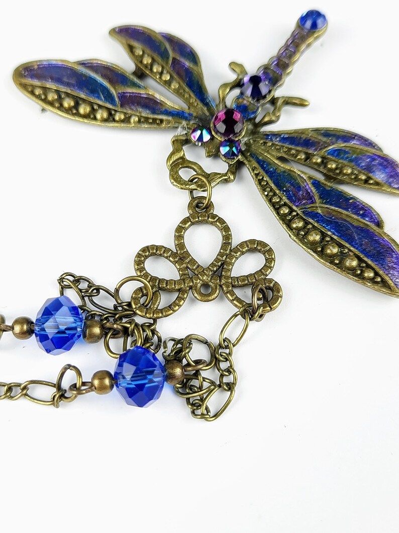 May include: A bronze dragonfly pendant with blue and purple iridescent wings, a decorative filigree charm, and two blue crystal beads hanging from a chain.
