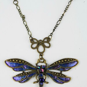 May include: A bronze necklace with a blue dragonfly pendant. The dragonfly has iridescent wings and a purple rhinestone body. The necklace has a chain with blue crystal beads.